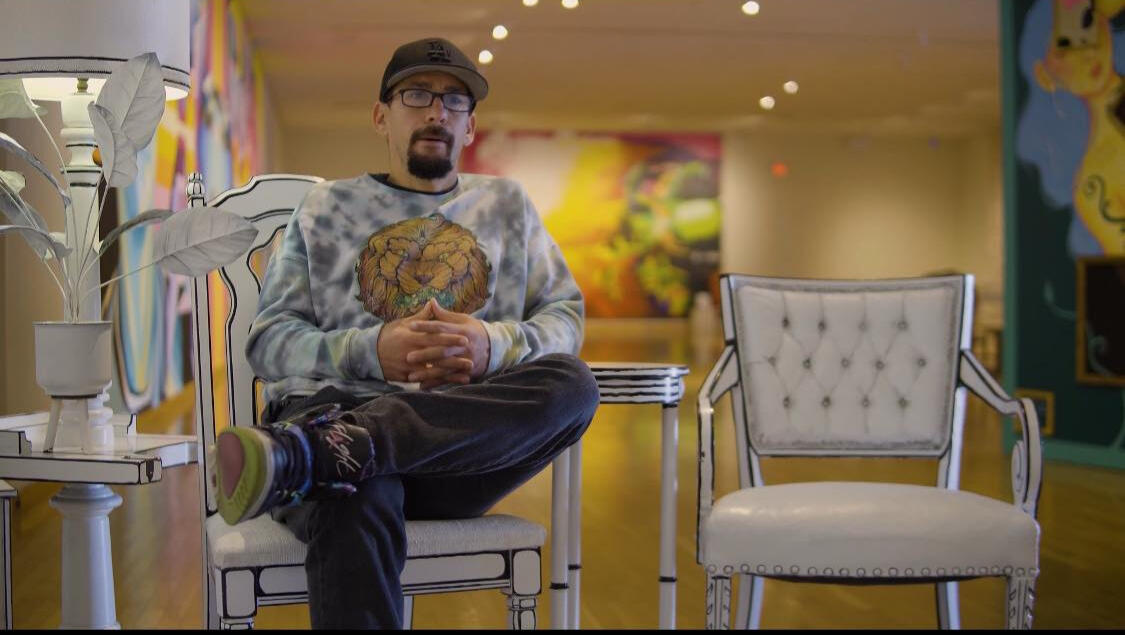 Lead commercial muralist Alex Van Galder giving a professional media interview seated in a custom chair during a fine art gallery exhibition by VanGalder Murals